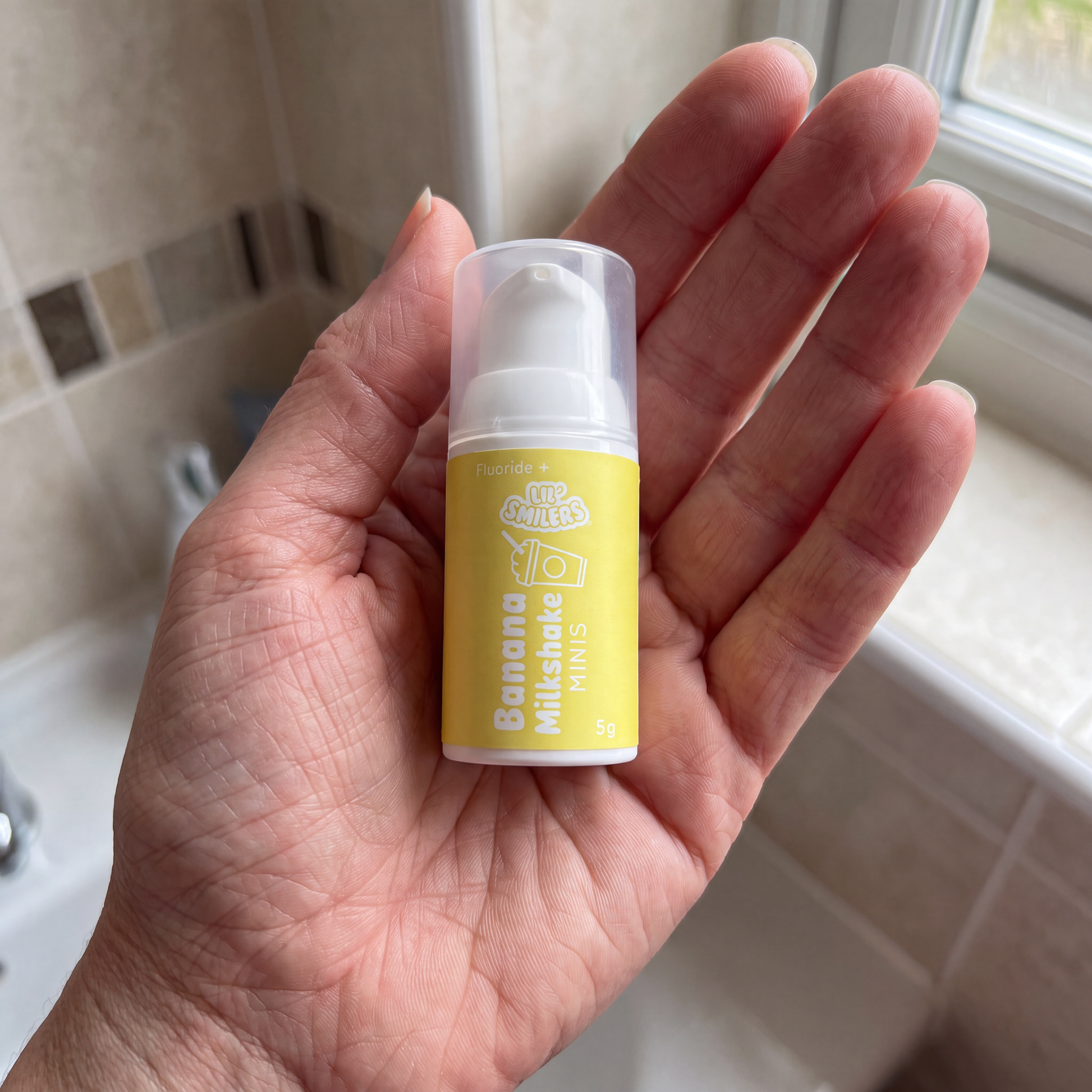 A hand holding a small bottle of Lil' Smilers Banana Milkshake MINIS toothpaste in a bathroom setting.