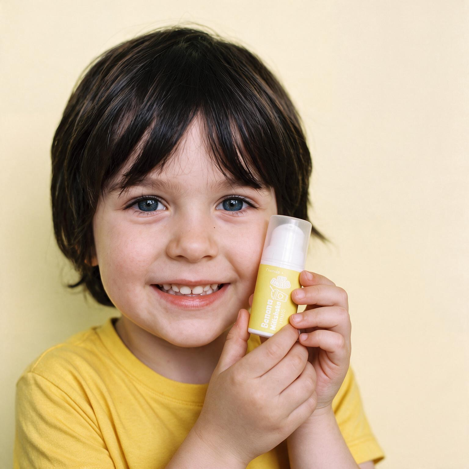A young child with dark hair and blue eyes smiles while holding a yellow cosmetic tube against a plain light beige background.