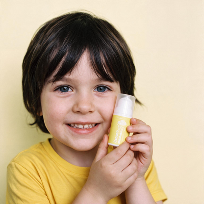 A young child with dark hair and blue eyes smiles while holding a yellow cosmetic tube against a plain light beige background.