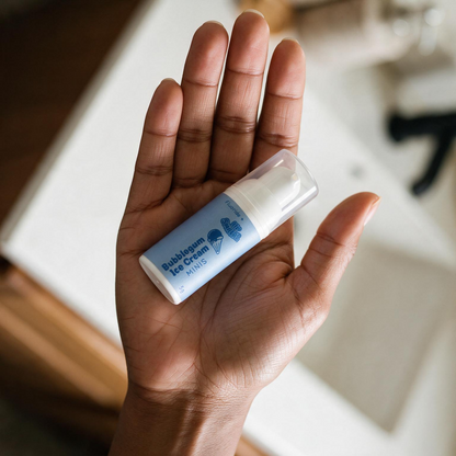 A hand holding a small bottle of  Lil' Smilers MINIS Bubblegum Ice Cream flavour toothpaste with a clear cap and light blue label in a bathroom setting.