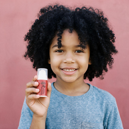A smiling young boy with curly black hair holds up a small red and white cosmetic tube against a solid dusty pink background.